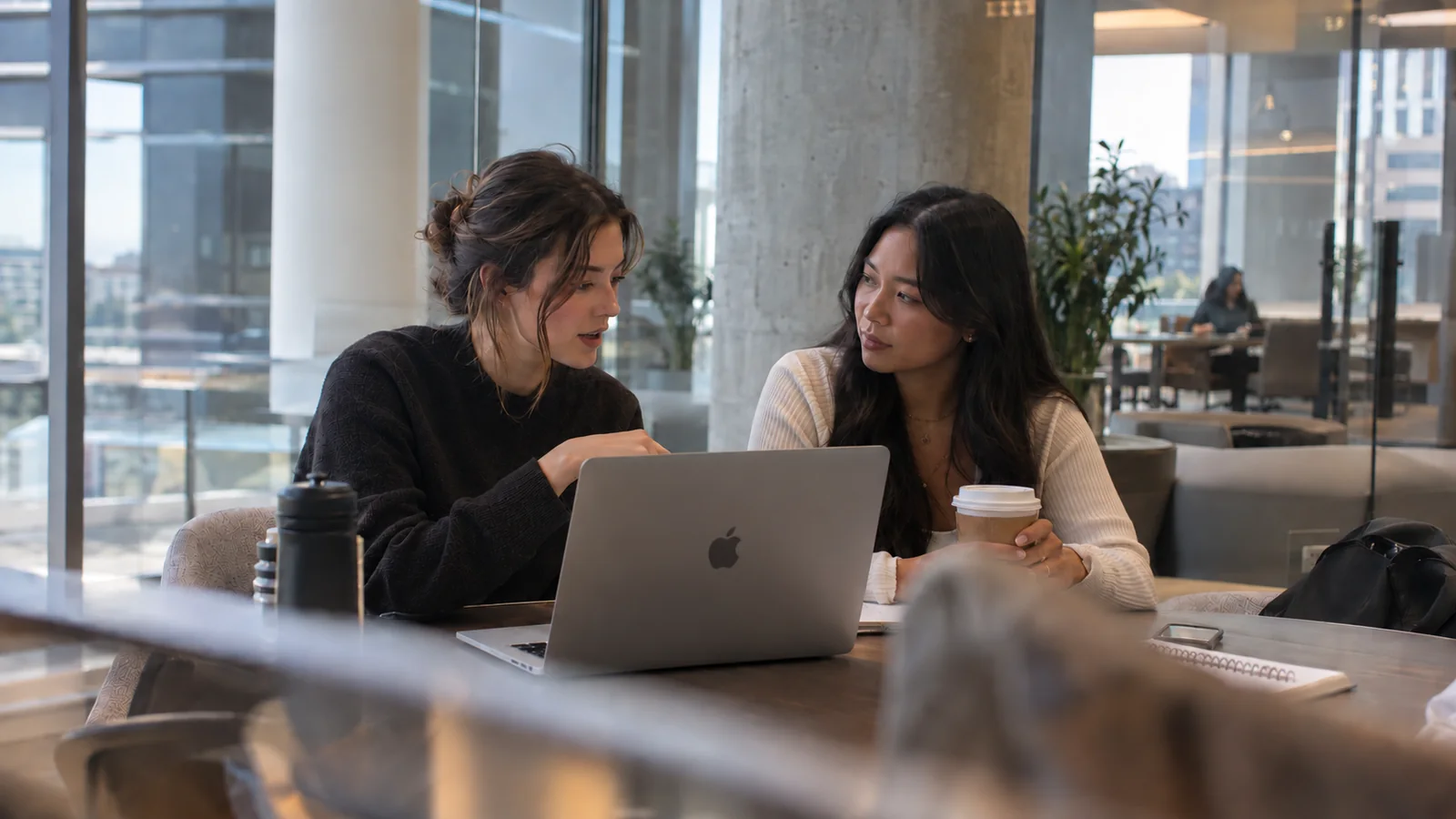 Two women seated at a table discussing information on a laptop, with a notebook and drinks nearby.